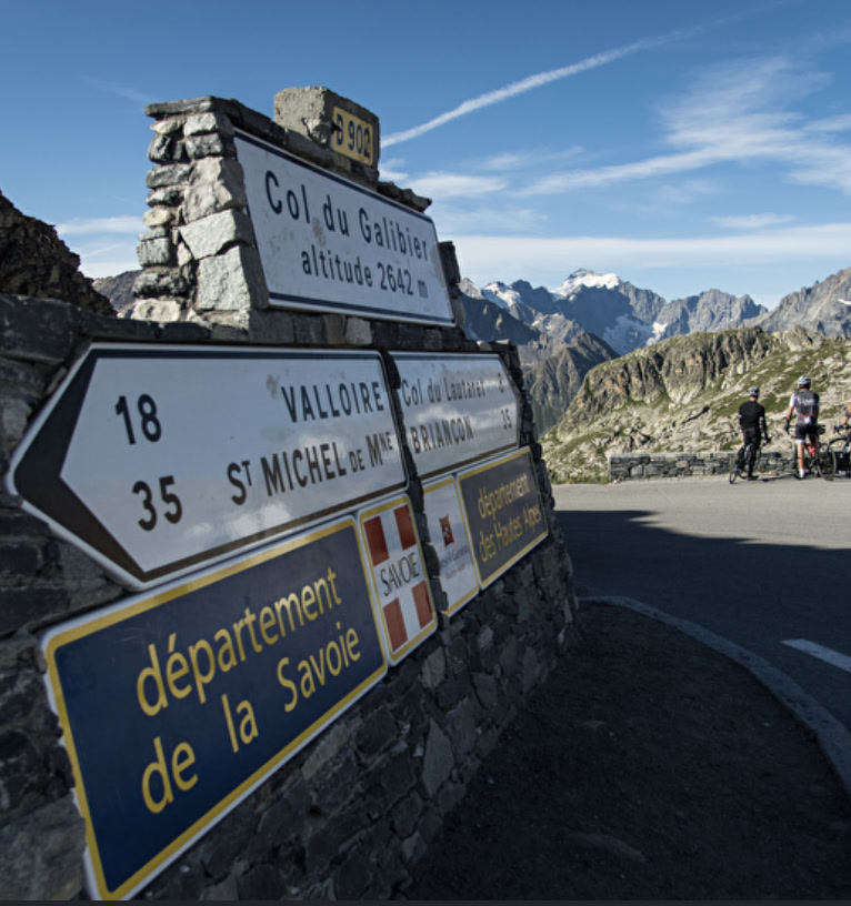 Summit signs at Col du Galibier altitude 2,642 m with alpine peaks and cyclists behind the stone wall